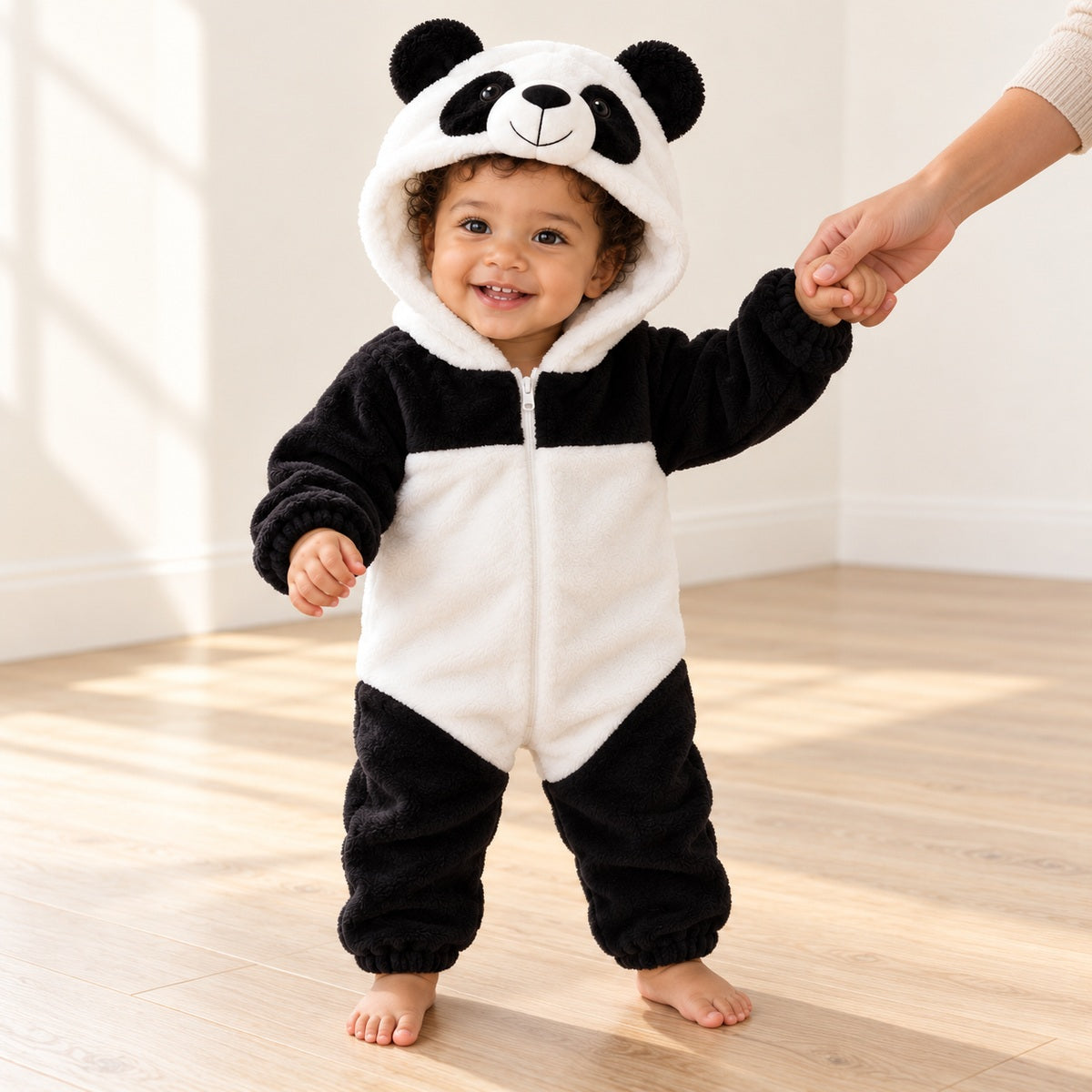 AI-generated toddler standing awake with an adult holding one hand, wearing the panda hooded romper in a neutral room.