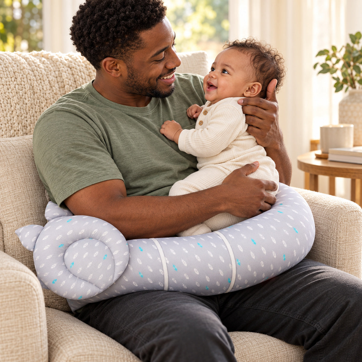 Parent holding an awake baby with the gray patterned nursing pillow across his lap at home